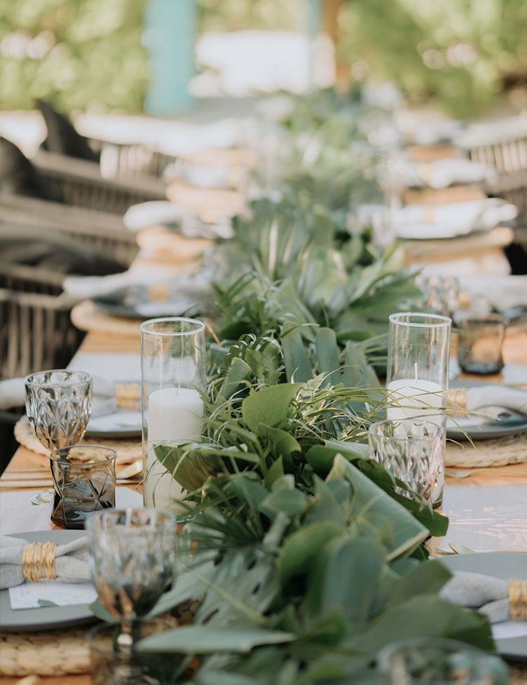 Long table set for an outdoor event with greenery centerpieces, candles, and place settings.