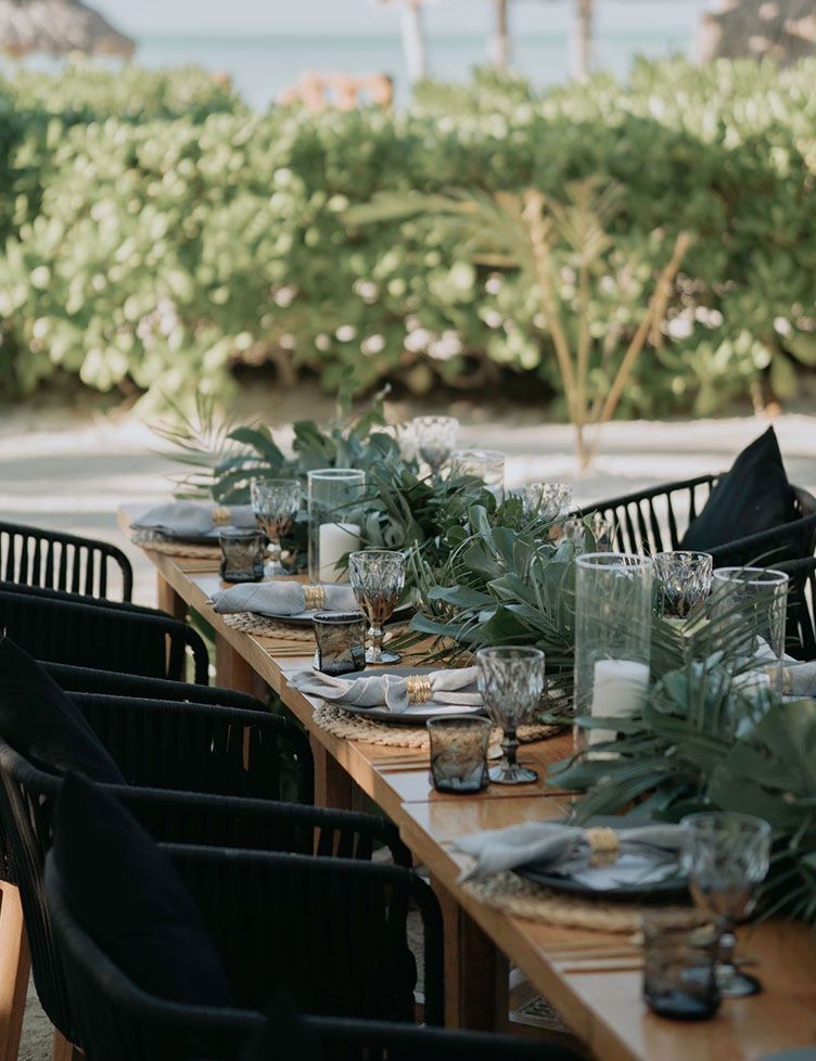 Long wooden table set for dining, black chairs, greenery centerpieces, beach setting.