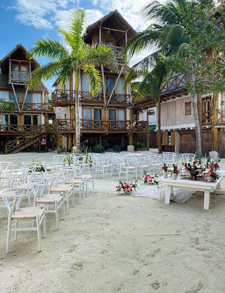 Beach wedding setup with white chairs and floral arrangements, wooden buildings in the background.