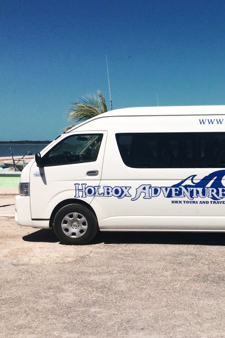 White Holbox Adventure van parked on a sunny day, near the water. Blue logo on the side.