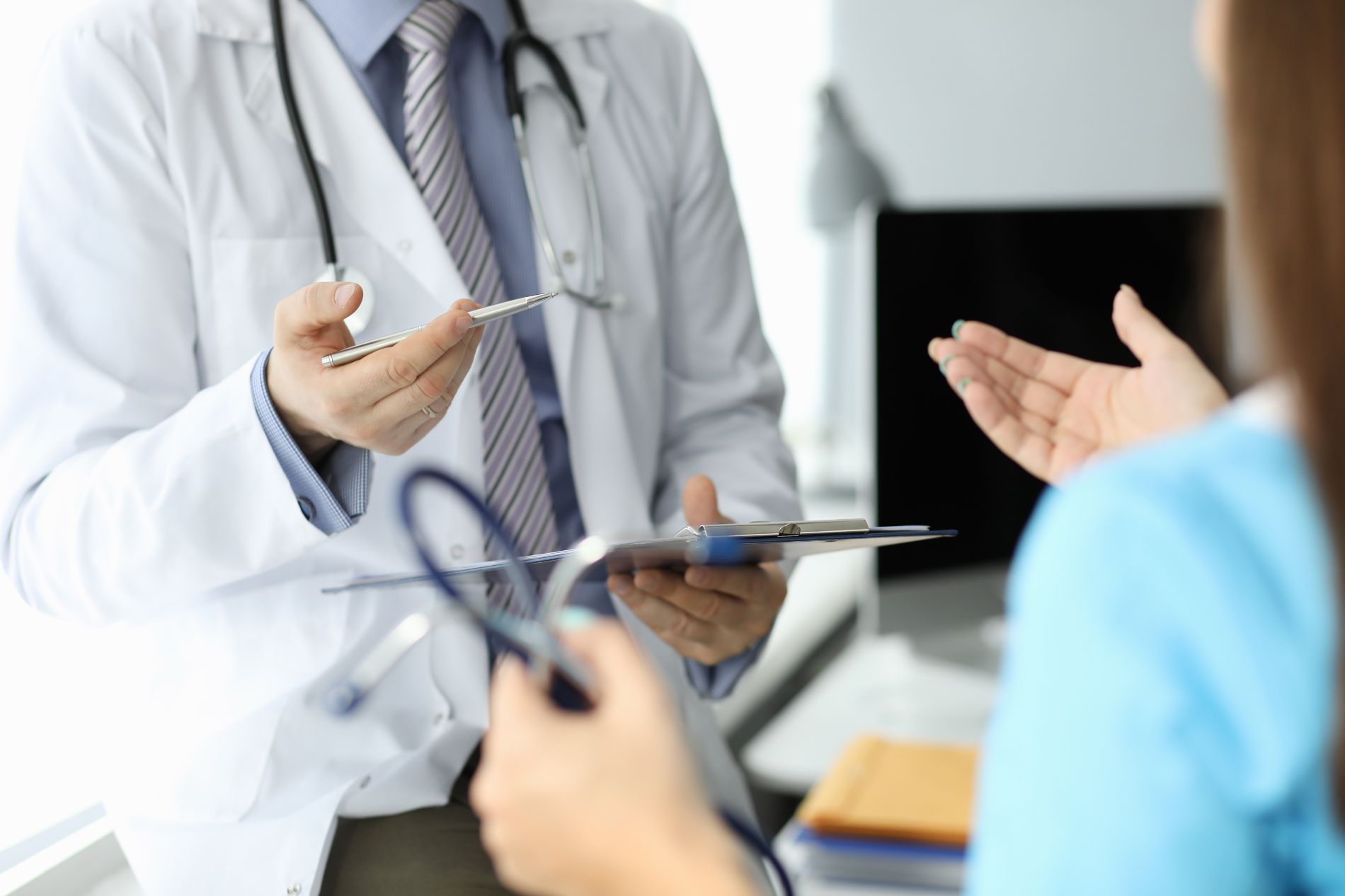 Un medico in camice bianco indica con una penna il paziente che sta gesticolando in un ambiente medico.
