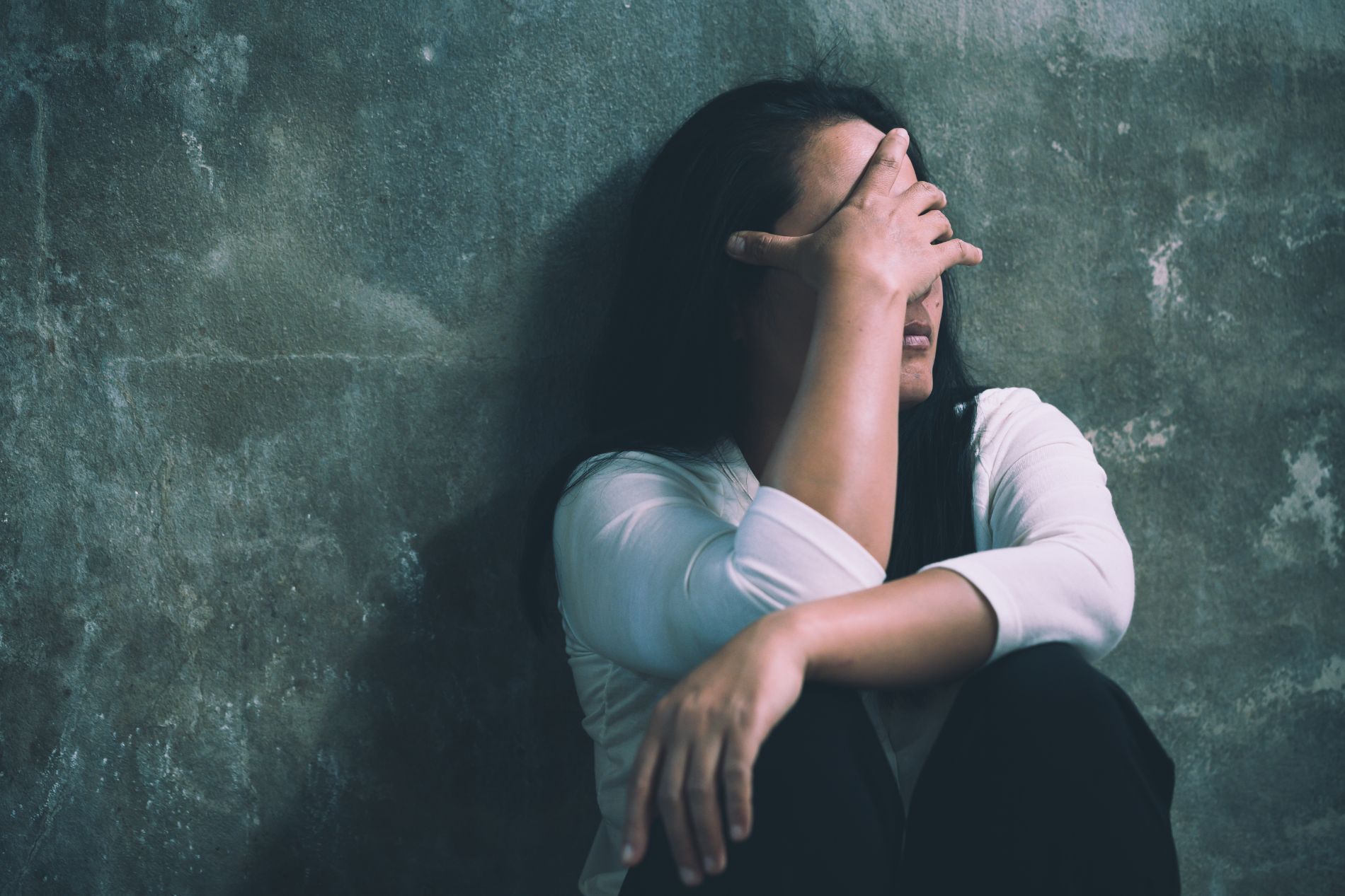 Una donna dai capelli scuri è seduta contro un muro grigio, si copre il viso con la mano e appare angosciata.