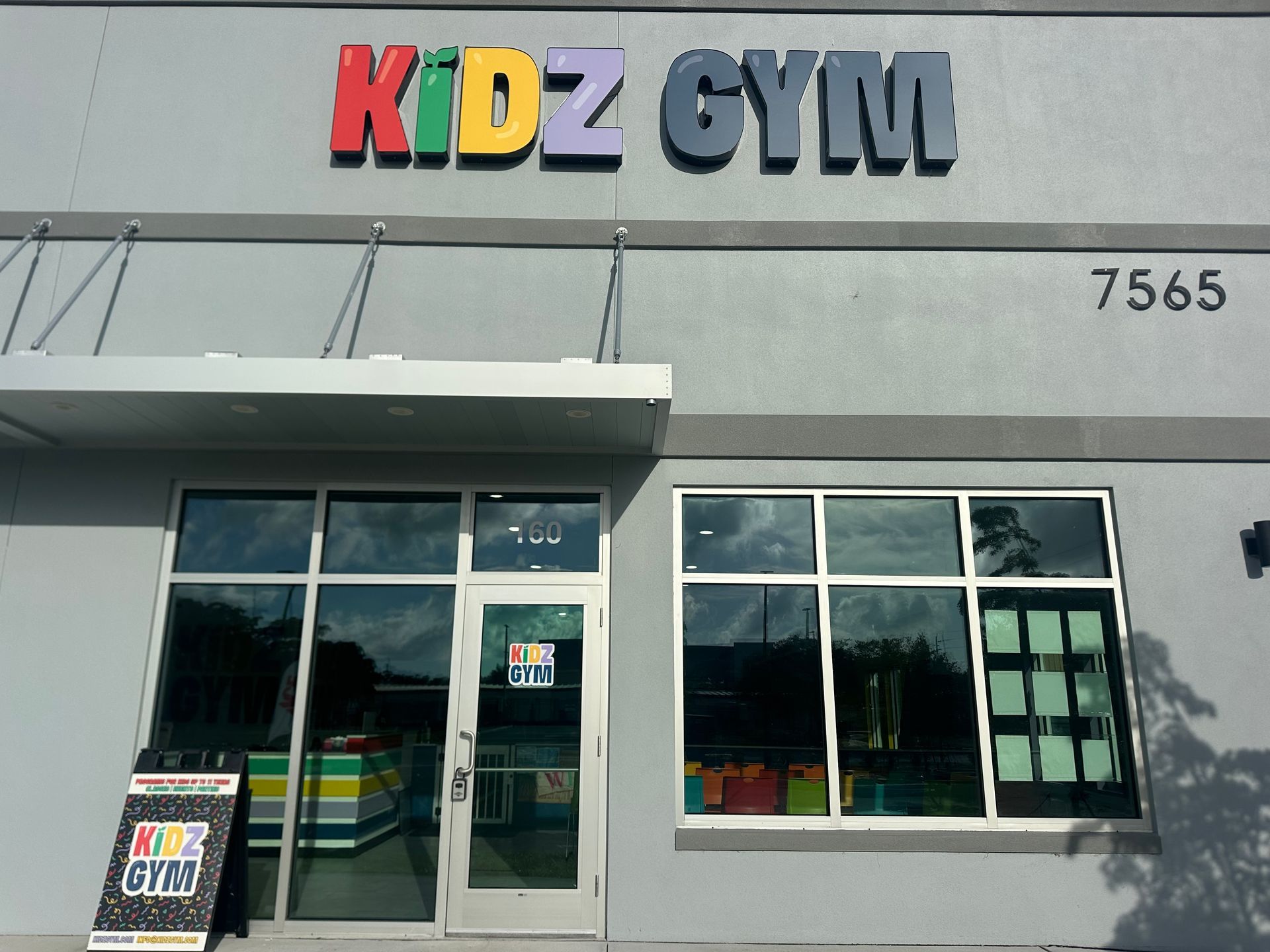 Kidz Gym exterior with colorful sign, awning, and windows showcasing gym equipment and activities.