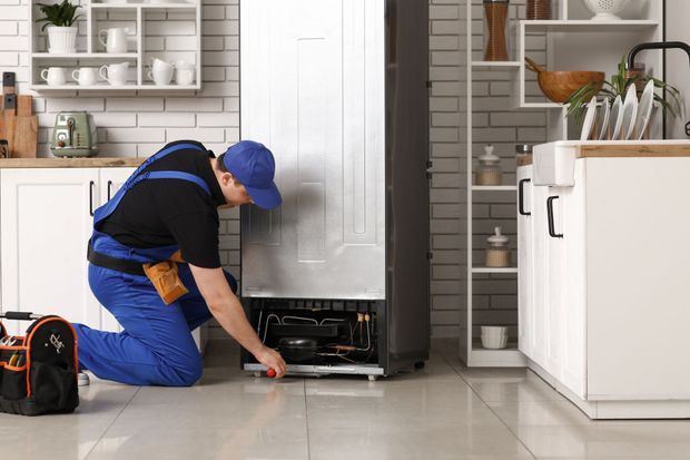 Technician in blue uniform kneels to repair an appliance in a bright kitchen.