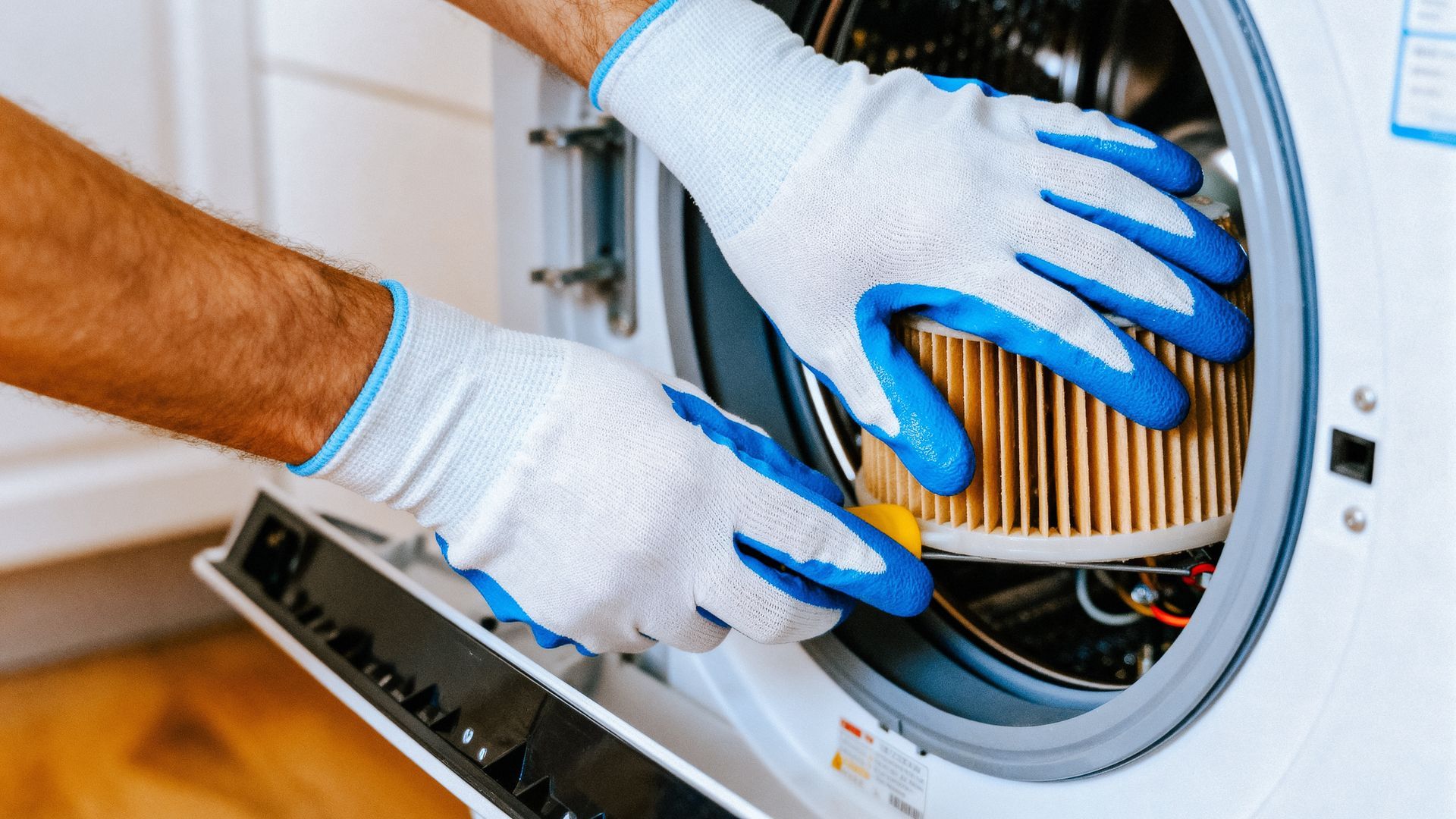 Gloved hands cleaning a washing machine’s filter, with lint and debris visible inside.
