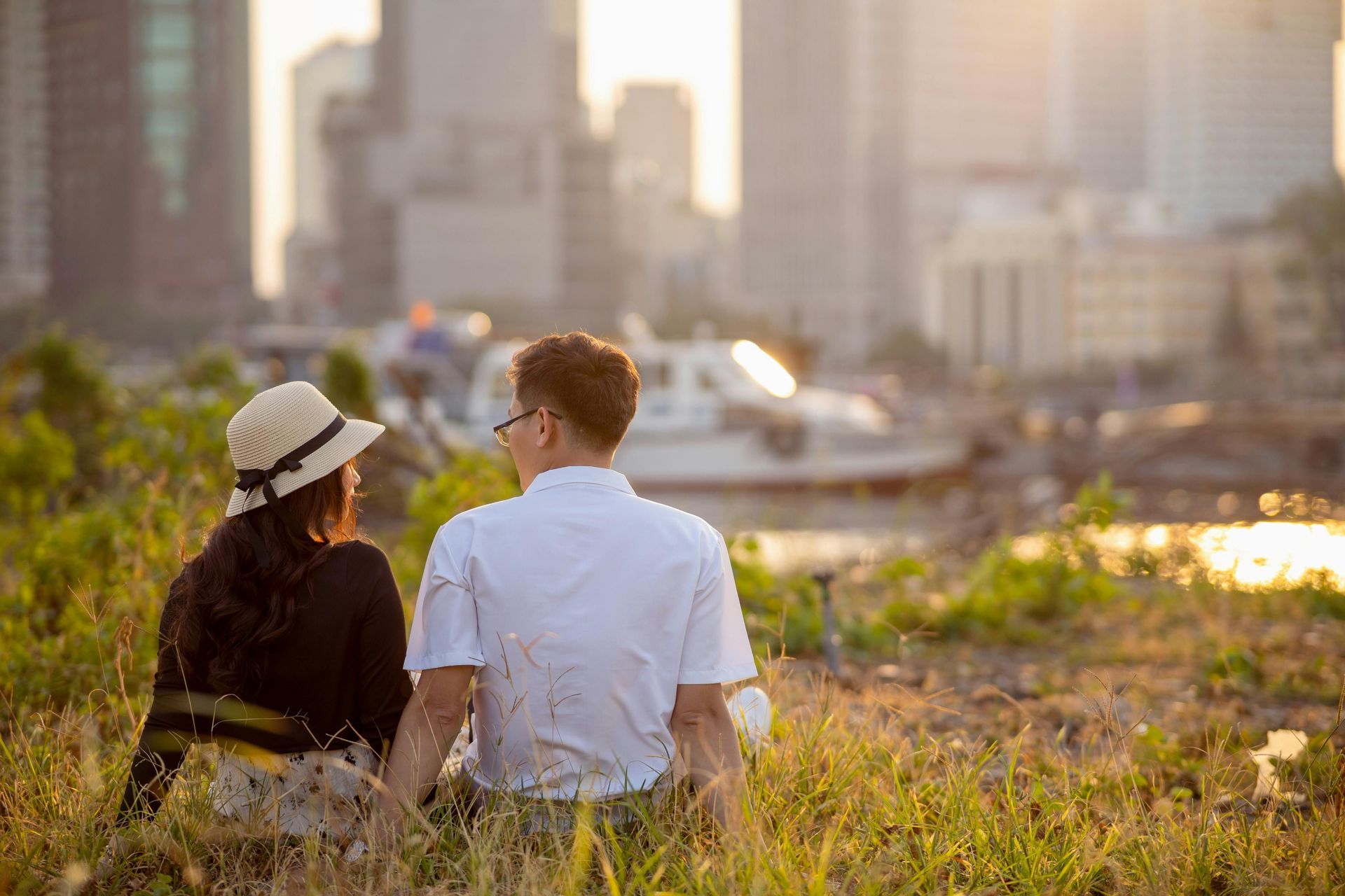 Two people sitting in a sunlit field, looking toward a city skyline at sunset.