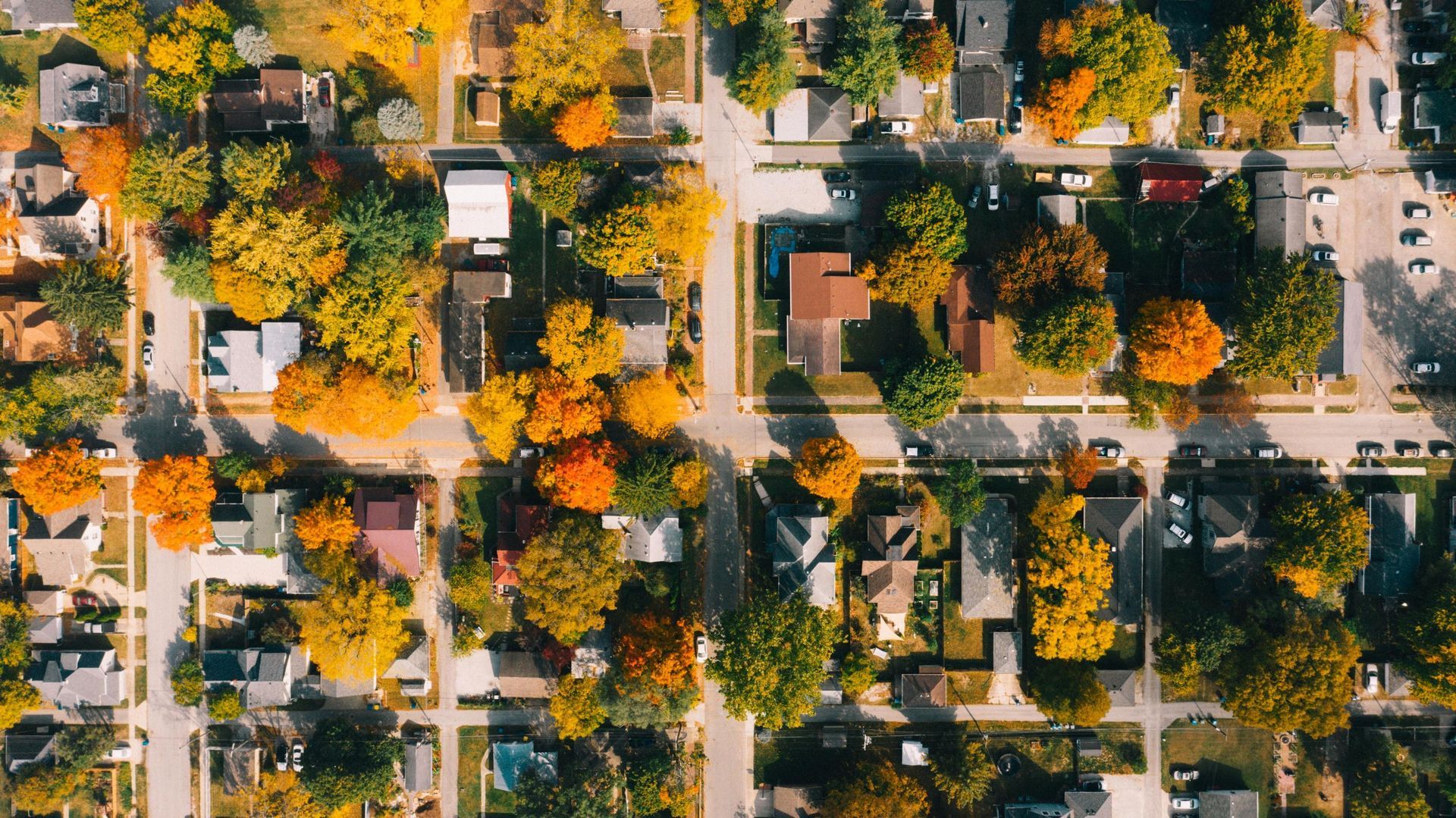 Aerial view of a neighborhood with tree-lined streets, houses, parked cars, and autumn foliage