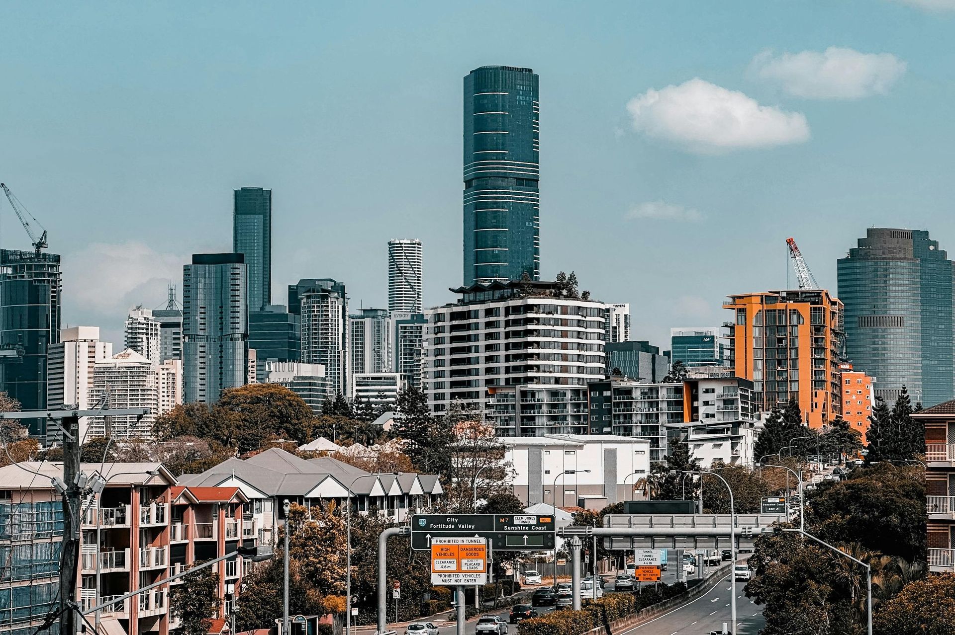 City skyline with modern high-rise buildings, including a tall turquoise tower, above low houses and trees under a blue sky.