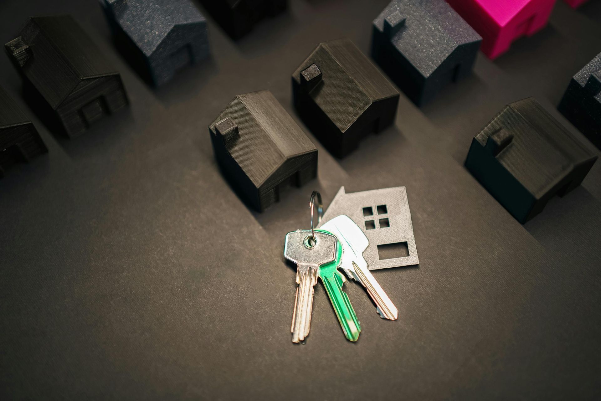Keys with a green keychain beside a small silver padlock on a dark tabletop, surrounded by miniature houses