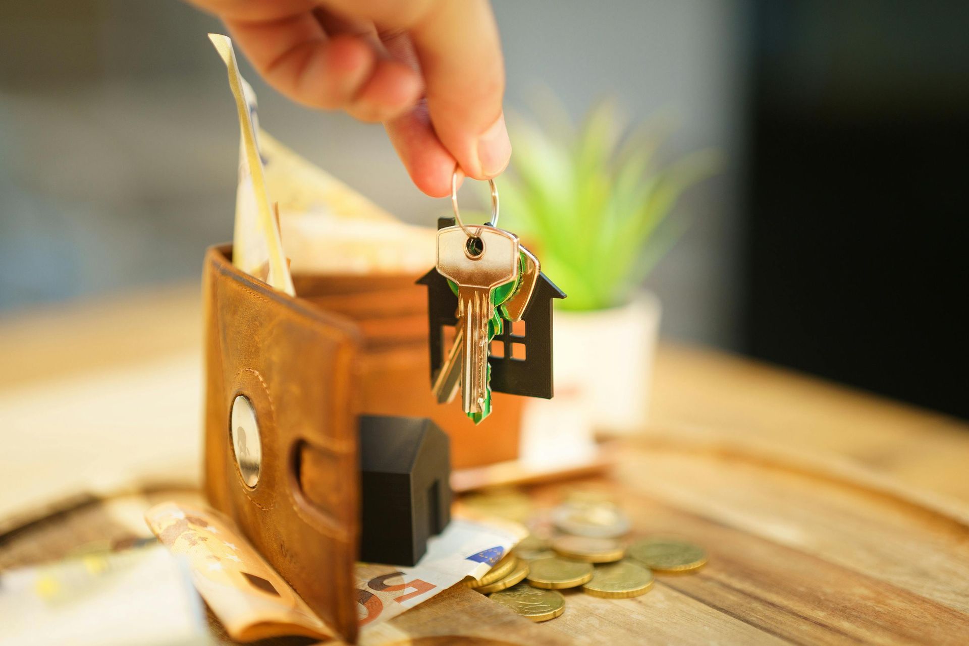 Hand placing house keys into a small wooden piggy bank beside scattered coins