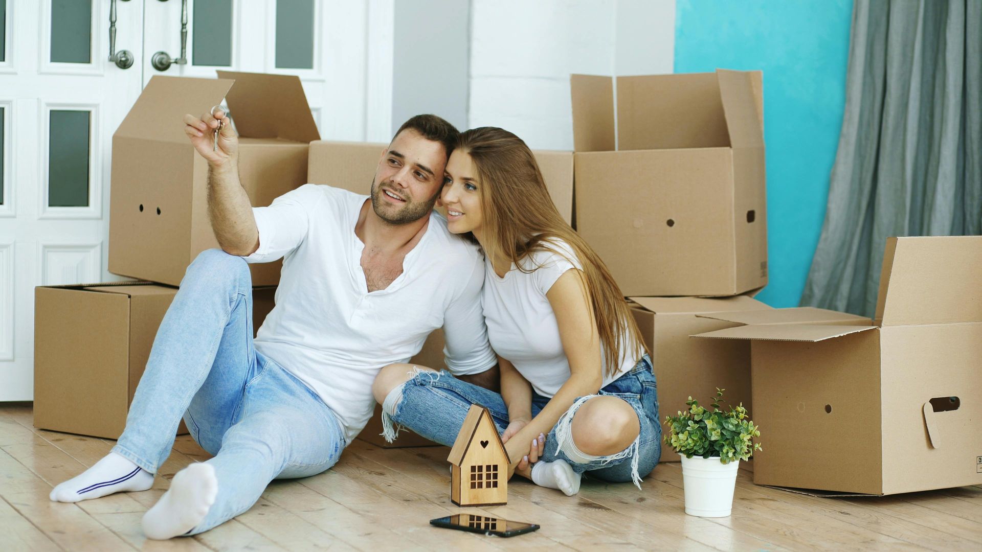 Couple sitting on the floor among moving boxes, taking a selfie in a bright new apartment.