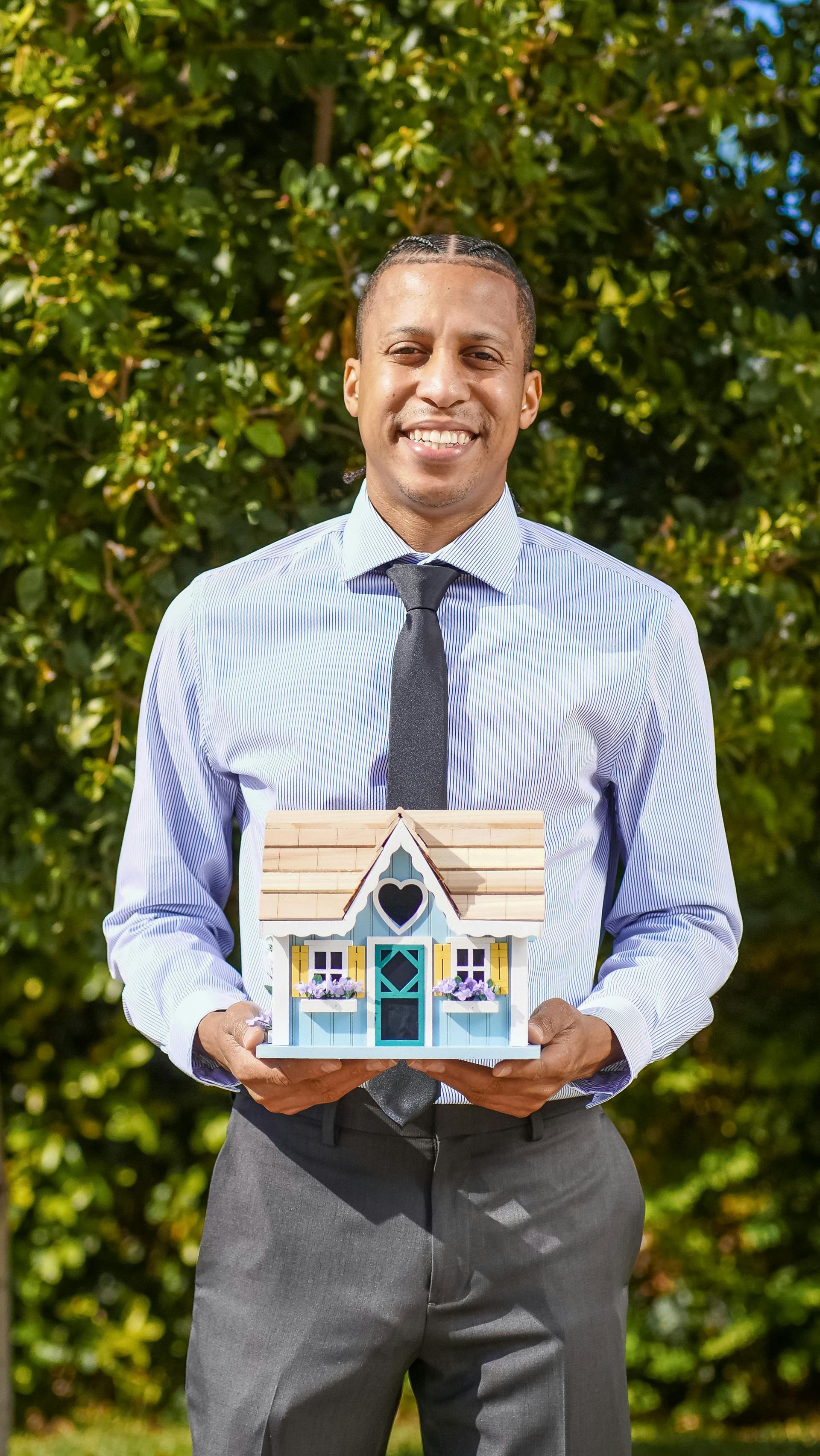 Man in striped shirt holding a small model house outdoors in front of green foliage