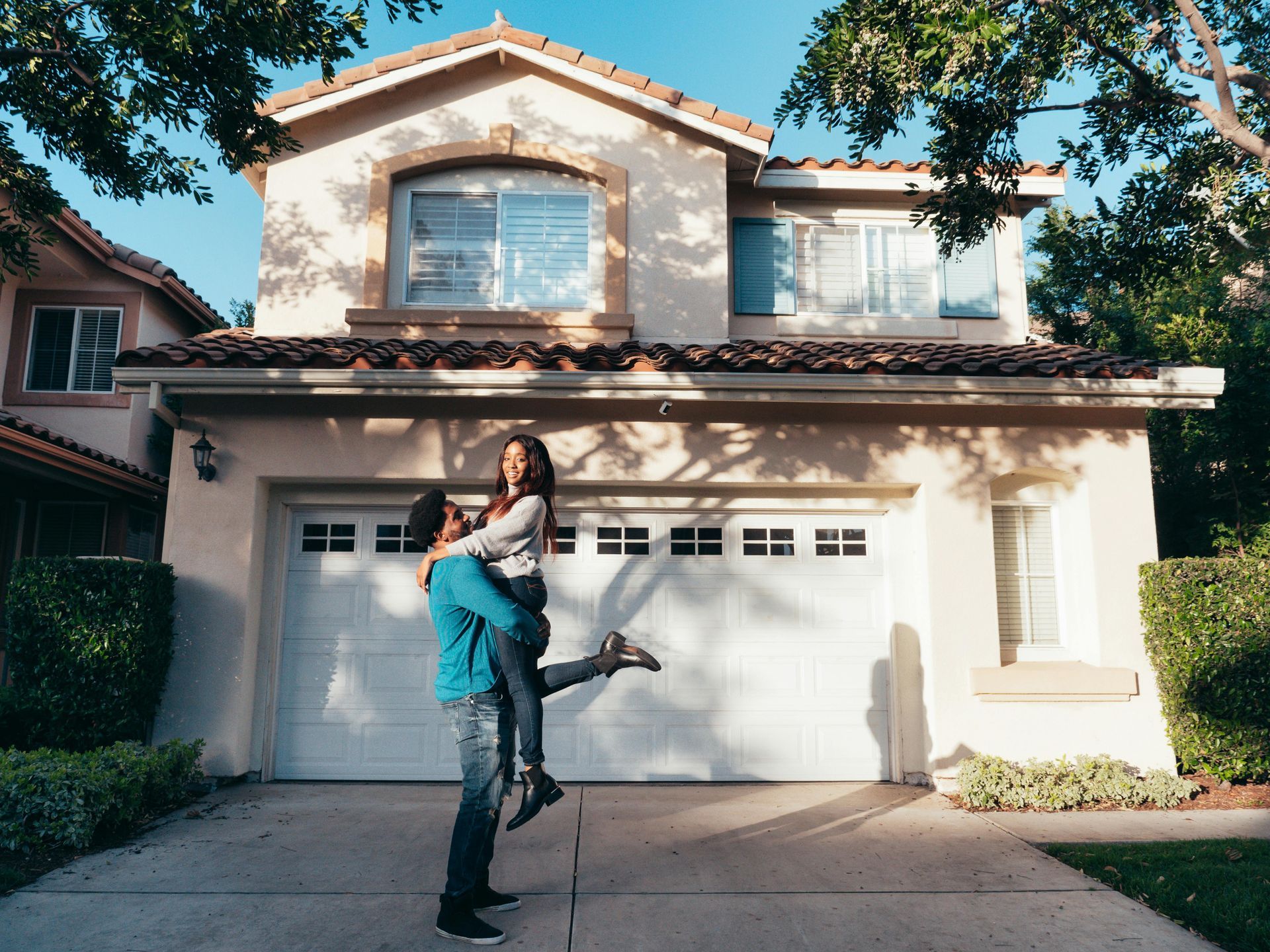 Man carrying a woman in front of a two-story suburban house with a white garage door