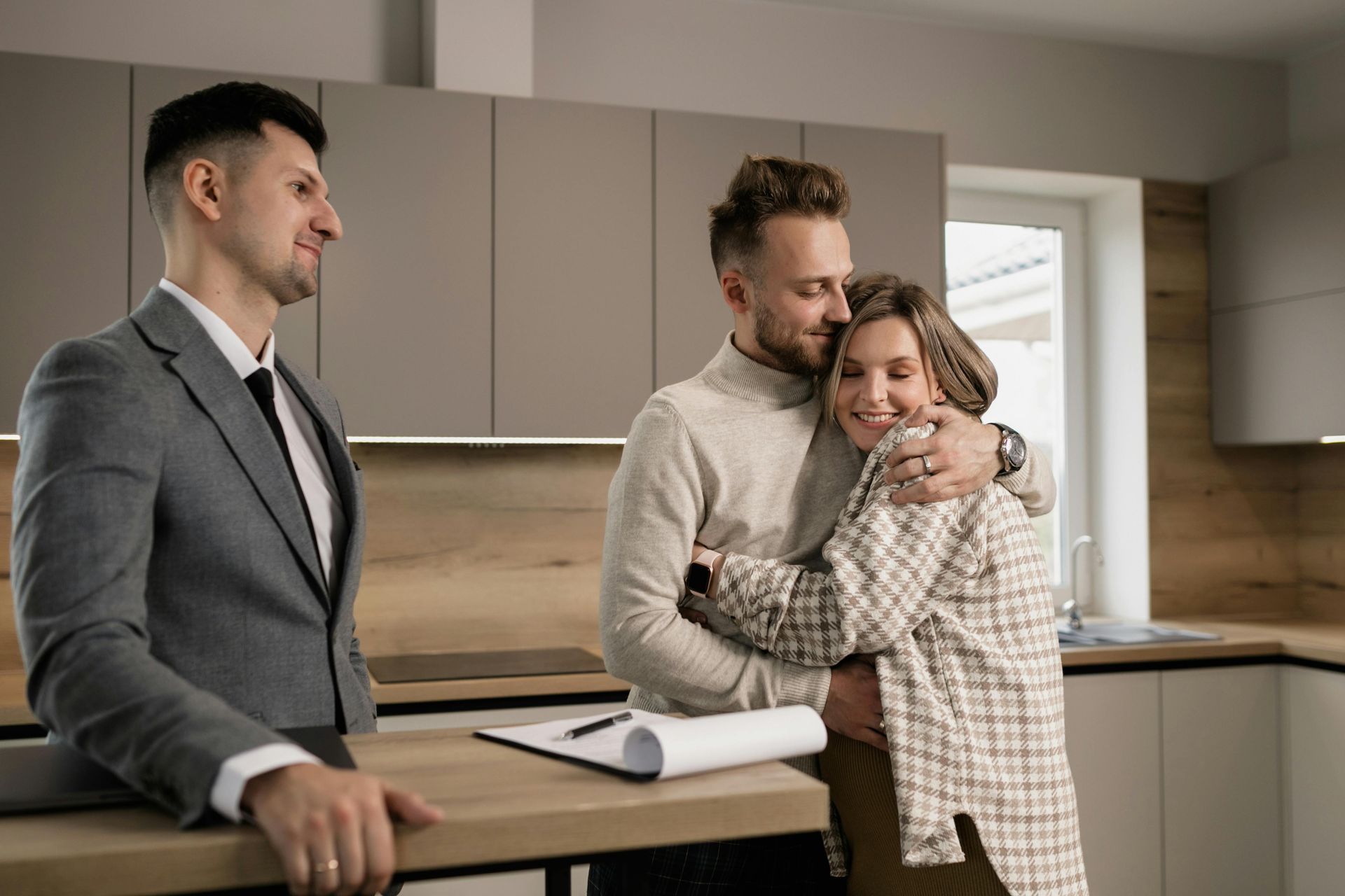 Three people in a modern kitchen, one couple hugging while a man stands nearby with a clipboard.
