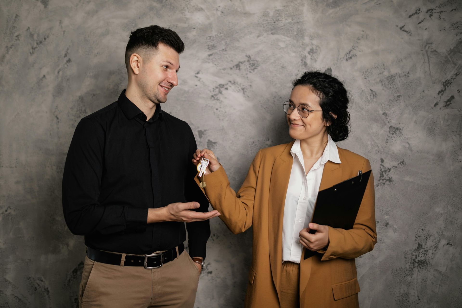 Two people in black and tan outfits smiling and holding a small object against a gray studio backdrop