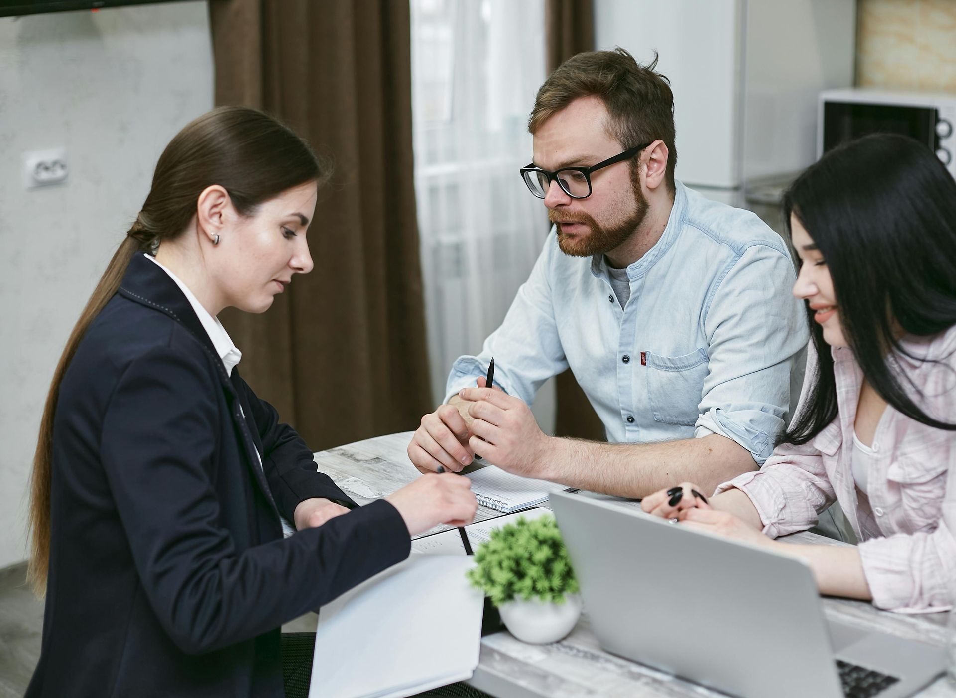 Three coworkers meeting around laptops, discussing documents at a table in a bright office