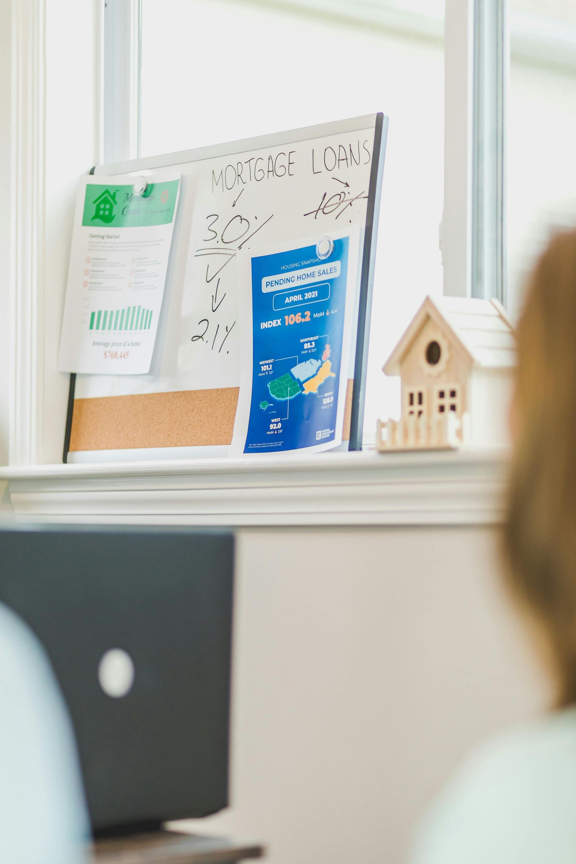 Office cubicle with a corkboard, blue brochure, small birdhouse decor, and a laptop on a desk.