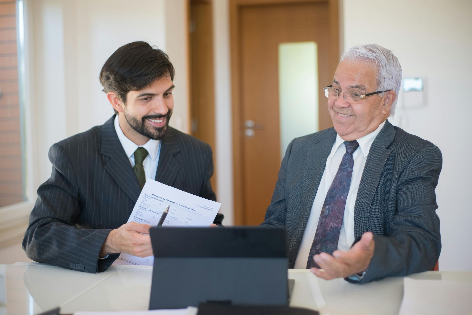 Two men in suits reviewing documents at a table with a laptop in an office