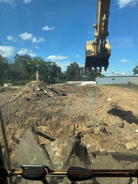 Dump truck dumping gravel onto a dirt road, under a blue sky. Excavator demolishing concrete and earth on a construction site; blue sky, trees in the background.