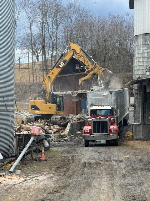 Yellow excavator working at a quarry, mountains in the background, dust clouds visible. Excavator demolishing building; semi-truck parked nearby. Debris scattered. Cloudy day.