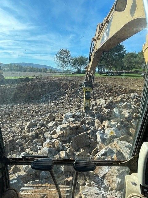 Yellow excavator digging earth on a construction site; blue sky in background. Excavator in action, breaking up rocks in a construction site with a blue sky overhead.