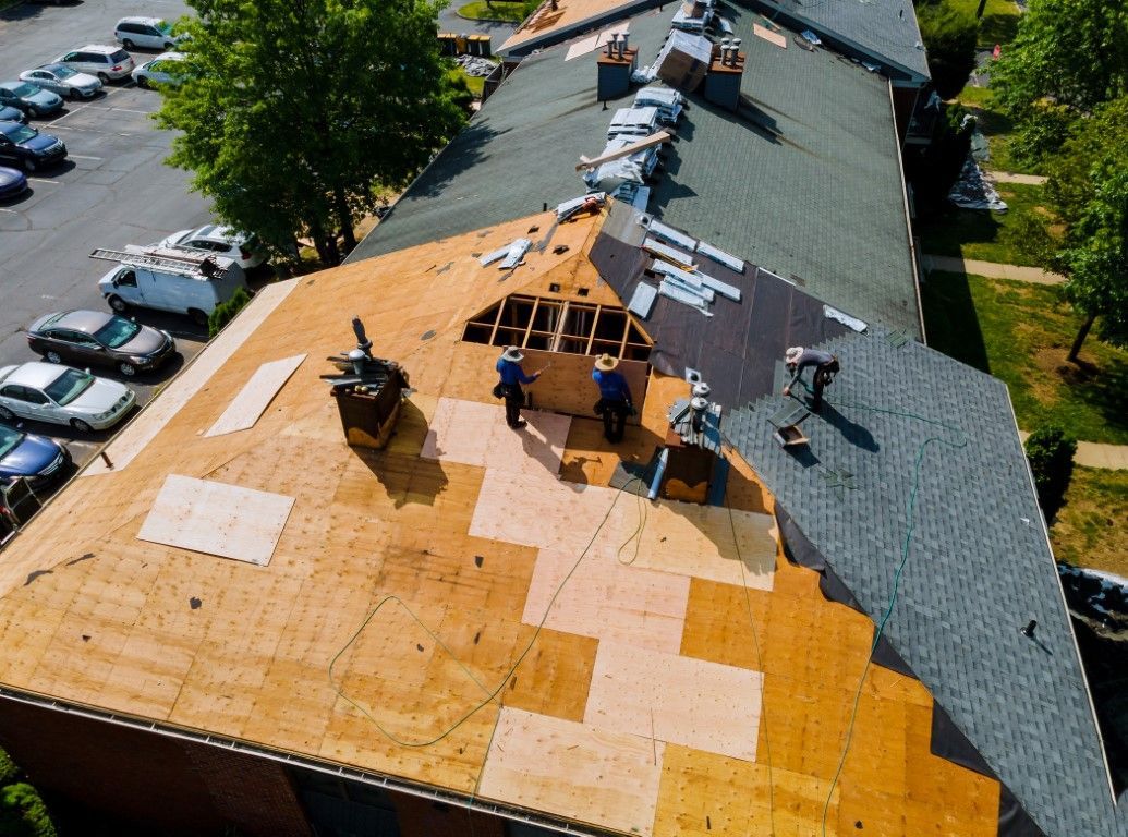 Roofers replacing shingles on a building. Aerial view shows exposed wood, new shingles, and parked cars.