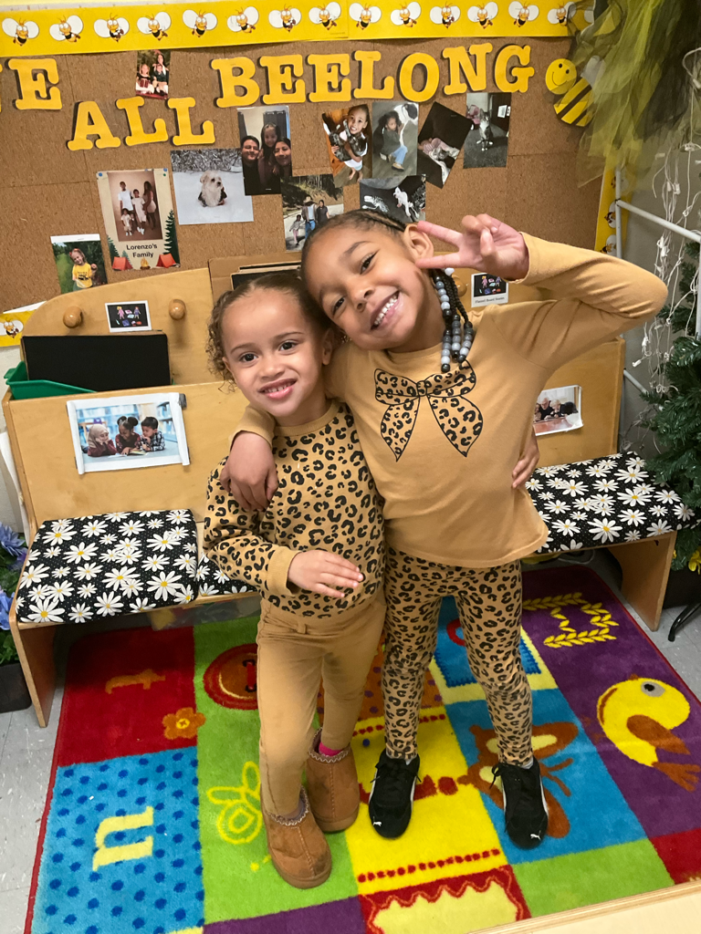 Two children posing on a colorful classroom rug in front of a “All Belong” bulletin board.