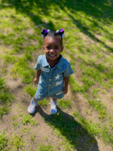 Smiling child in a denim dress standing on grassy ground in sunlight