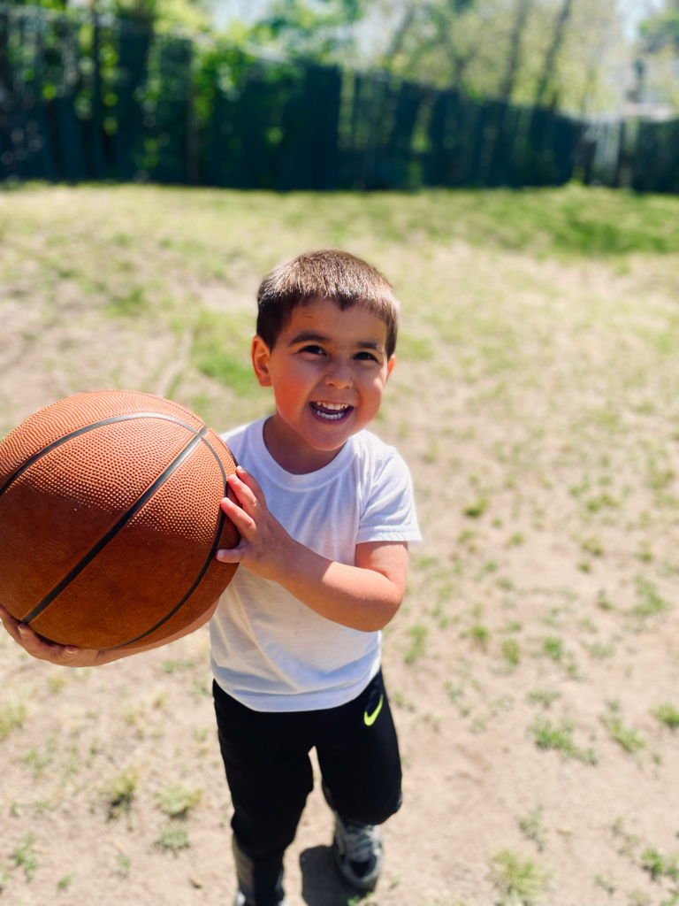 Child holding a basketball outdoors on a sunny grassy field, smiling at the camera