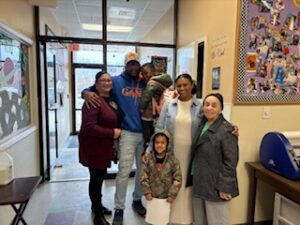 Five people posing in a hallway, smiling near an office entrance and bulletin board.
