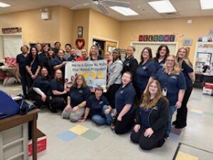 Group of staff in a classroom, posing together around a sign, smiling for a group photo.