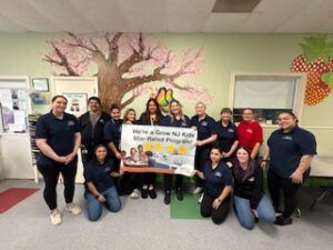 Group photo of smiling staff holding a banner in a classroom with a painted tree mural and colorful decorations