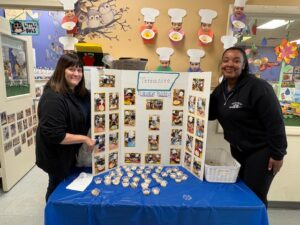 Two people stand by a blue-covered display board with photos and crafts in a classroom.