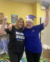 Two people smiling in a classroom, wearing blue and black shirts and graduation-style hats, holding small items aloft