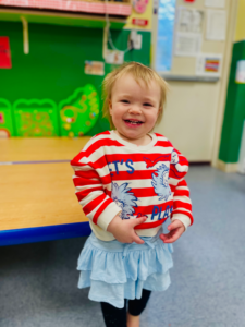 Smiling child in red-and-white striped shirt and blue skirt standing in a classroom