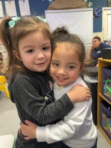 Two children hugging and smiling indoors, with classroom shelves and a seated adult in the background.