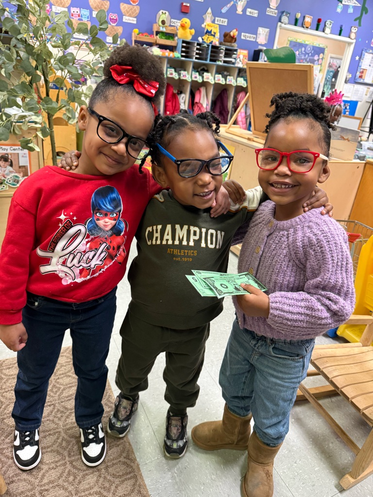 Three children standing together indoors, smiling and holding up a dollar bill.