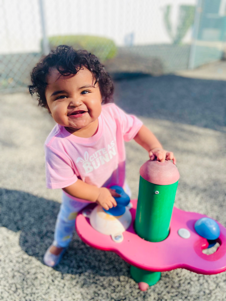 Smiling toddler in pink shirt pushing a pink ride-on toy outdoors