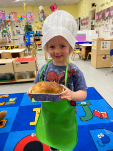 Child in a chef hat and green apron holding a baked dish in a classroom.
