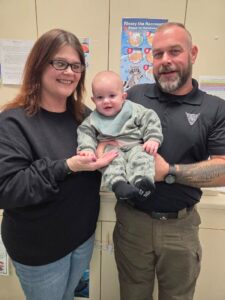 Three people standing indoors, holding a baby, with a colorful poster in the background.