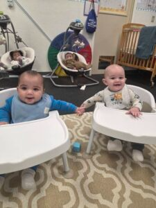 Two babies sitting in white high chairs at a home, smiling and holding hands.
