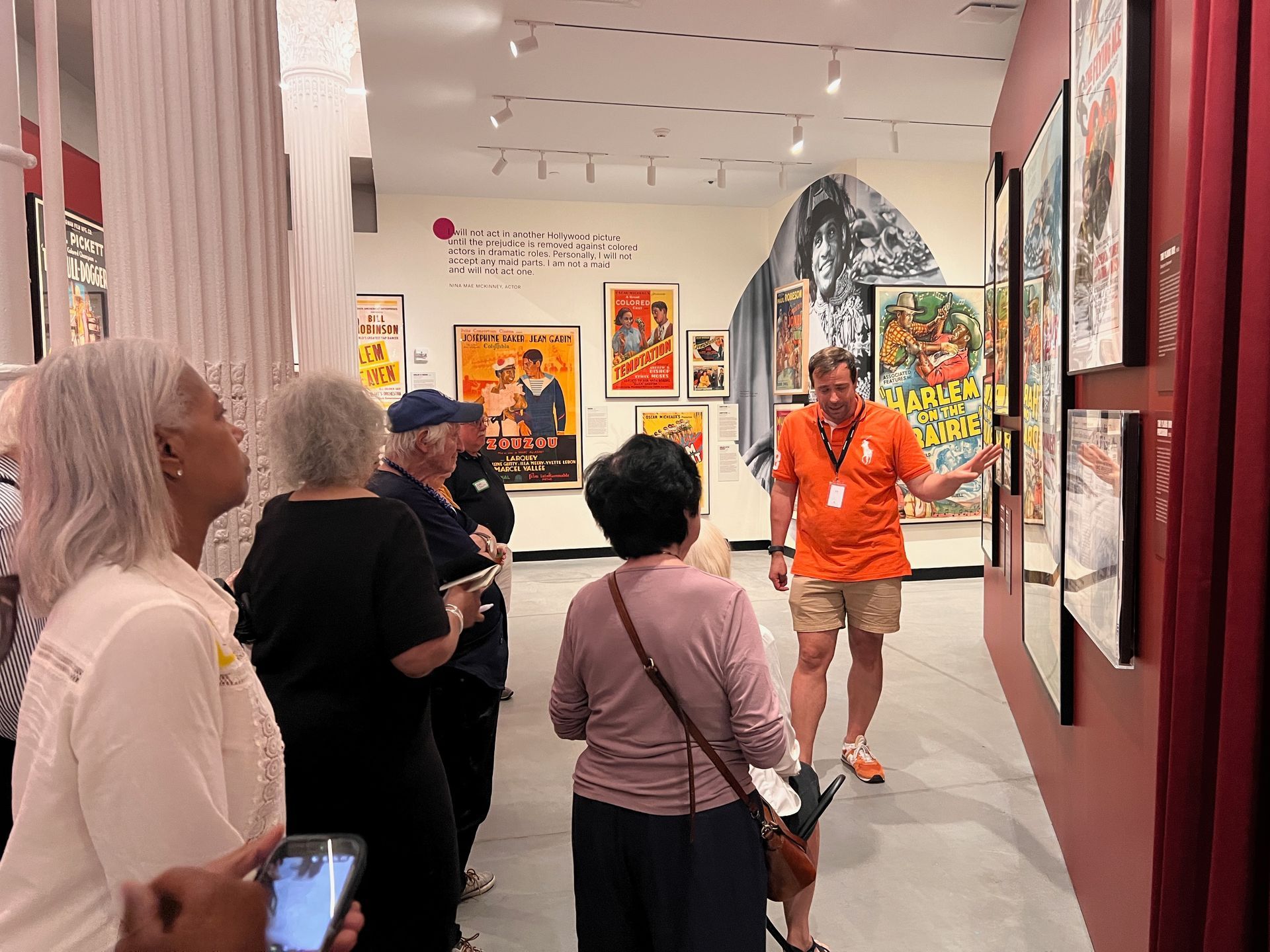 People viewing colorful paintings in a bright art gallery, with a guide in an orange shirt speaking.