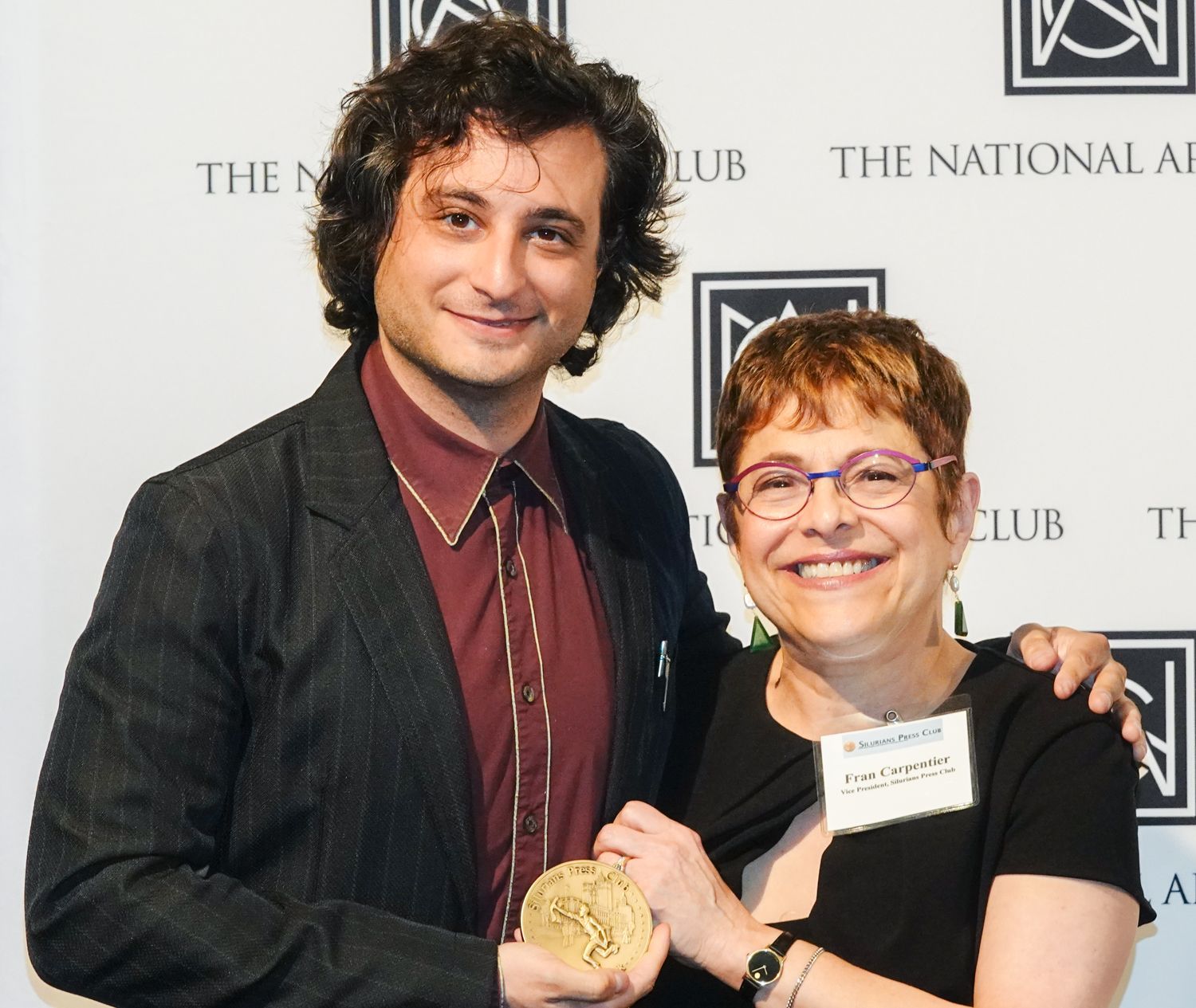 Man in a suit jacket with curly hair presents a gold medal to a woman in glasses smiling at The National Arts Club.