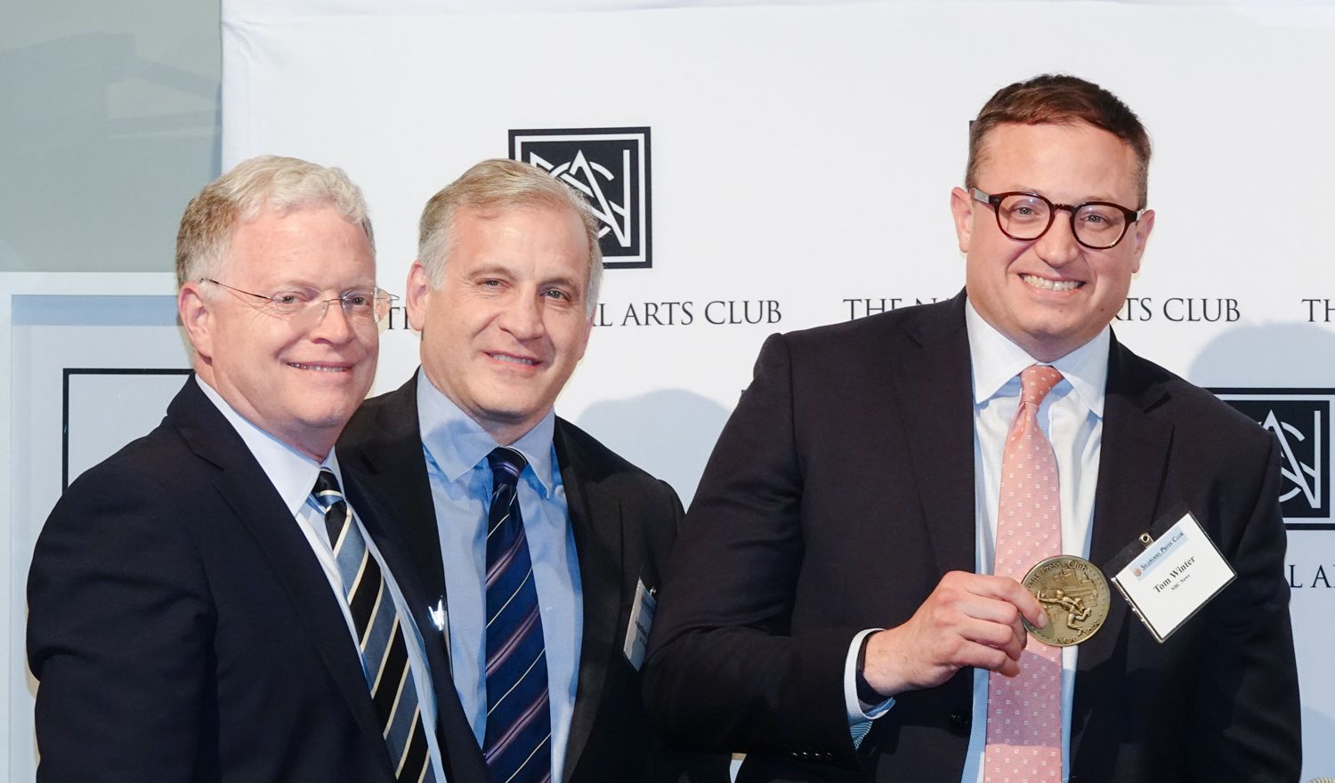 Three men in suits; one holding a gold medal at The Arts Club.