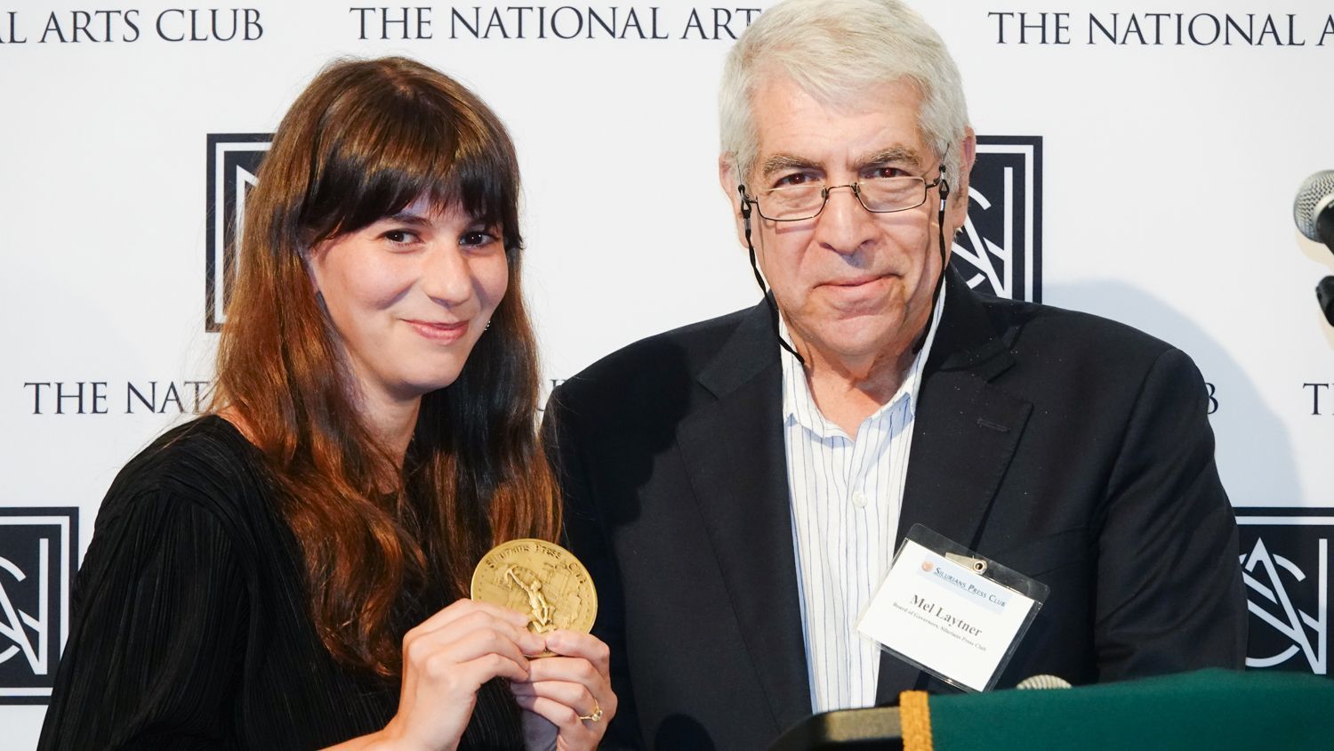 Woman holds gold medal next to older man at National Arts Club. Both smile.