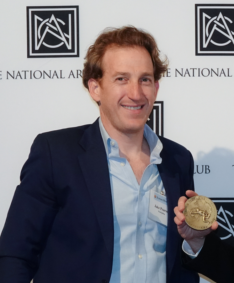 Man in blue blazer holds a gold medal, smiling. The setting appears to be a formal event.