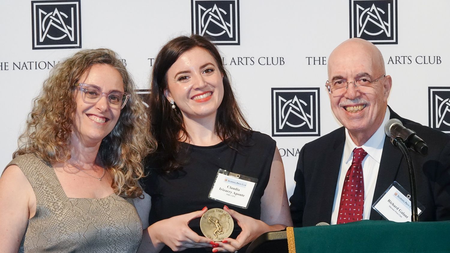 Three people at podium, woman in black dress holding a gold medal, two others smiling beside her.