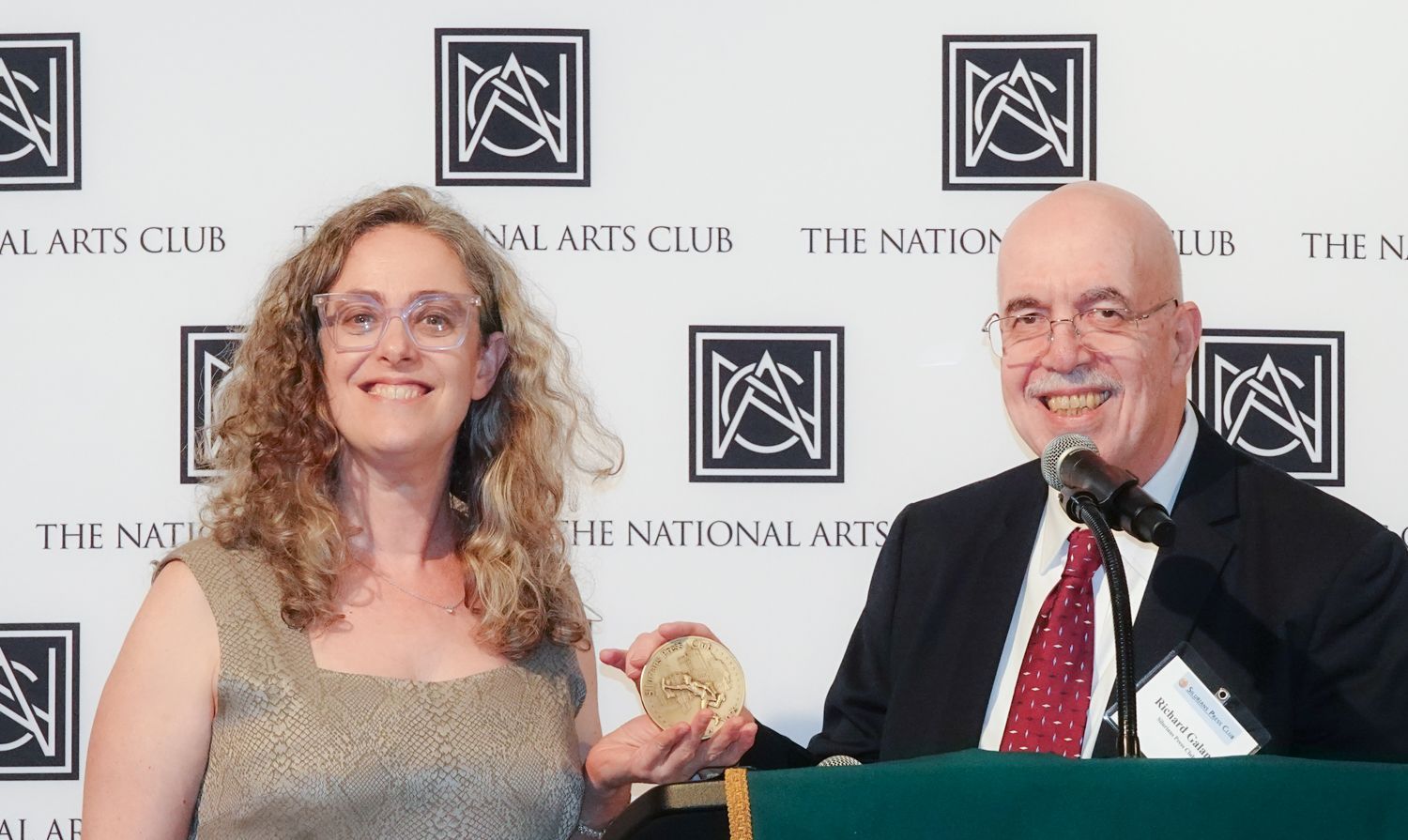 A woman and a man at a podium, holding a gold medal. They are at The National Arts Club.
