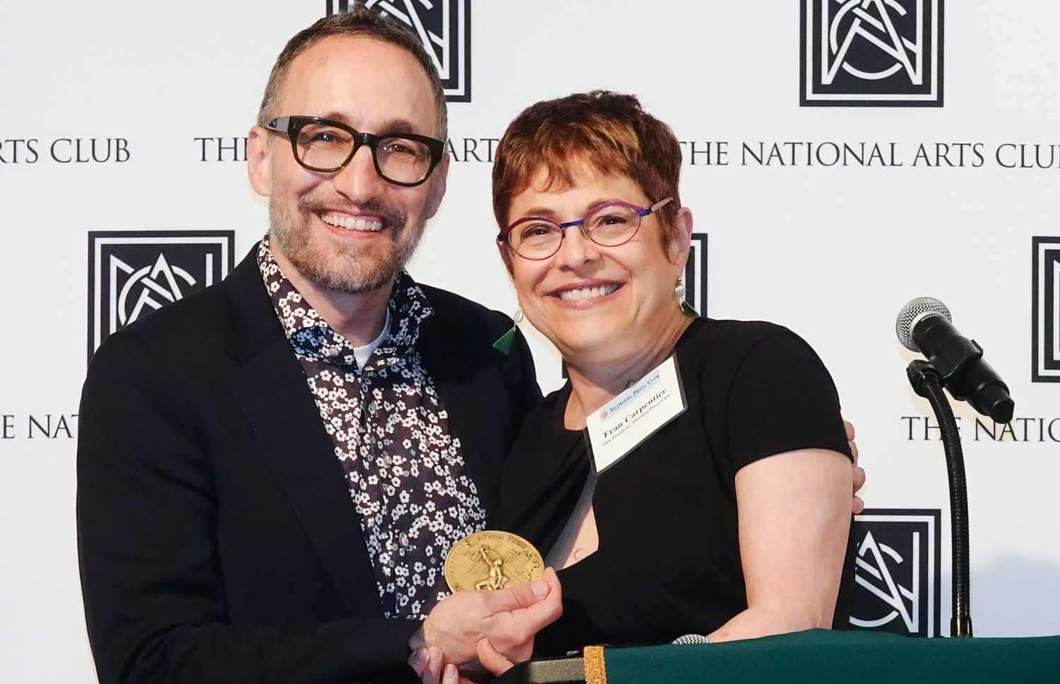 Two people smiling, one holding a gold medal. They are in front of a podium with
