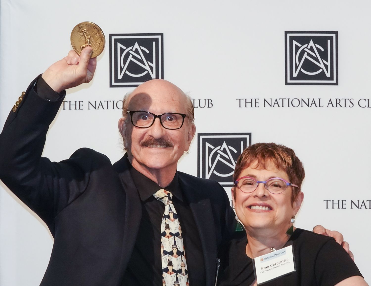Man holding gold medal, smiling, with woman next to him. National Arts Club backdrop.